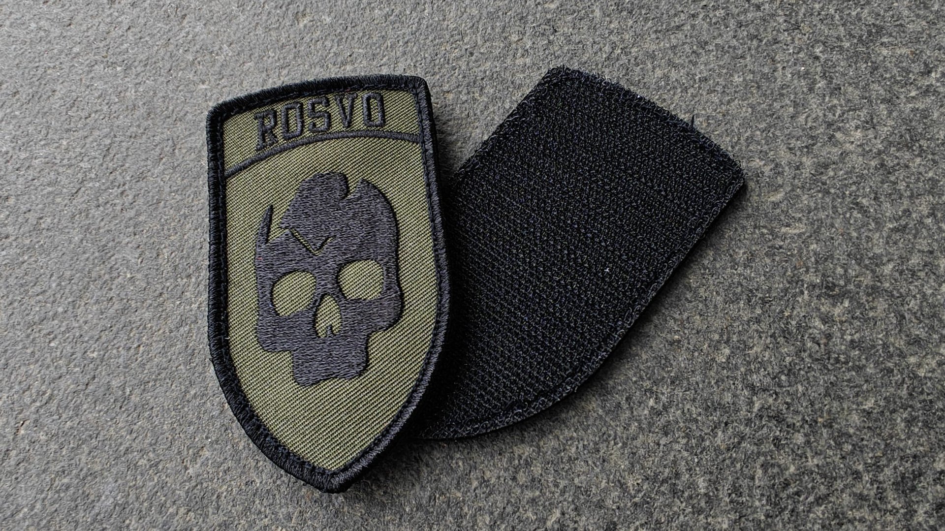 A military-style patch featuring a skull logo and the text ROSGVD on a gray shield, lying on a textured surface.