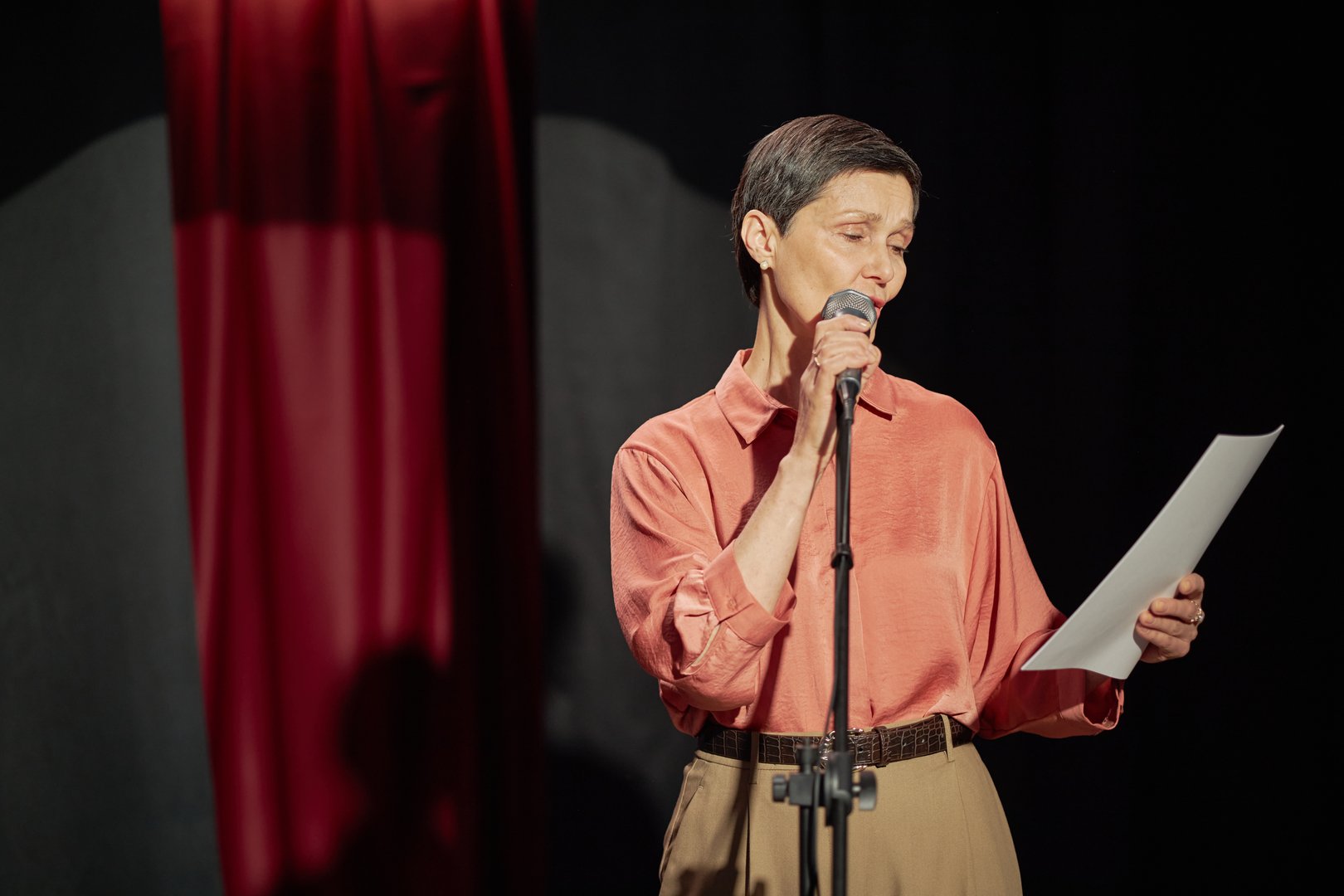 Middle aged Caucasian woman standing on stage holding microphone and reading from paper during talent competition, focused expression, short hair, stage curtain in background