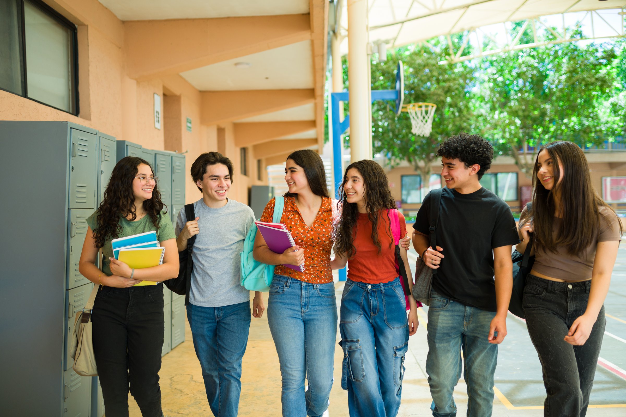Group of cheerful latin teenage students walking together in school hallway and carrying backpacks and notebooks