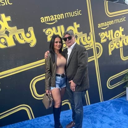 A man and woman pose together on a blue carpet at an Amazon Music event with a 24k party theme.