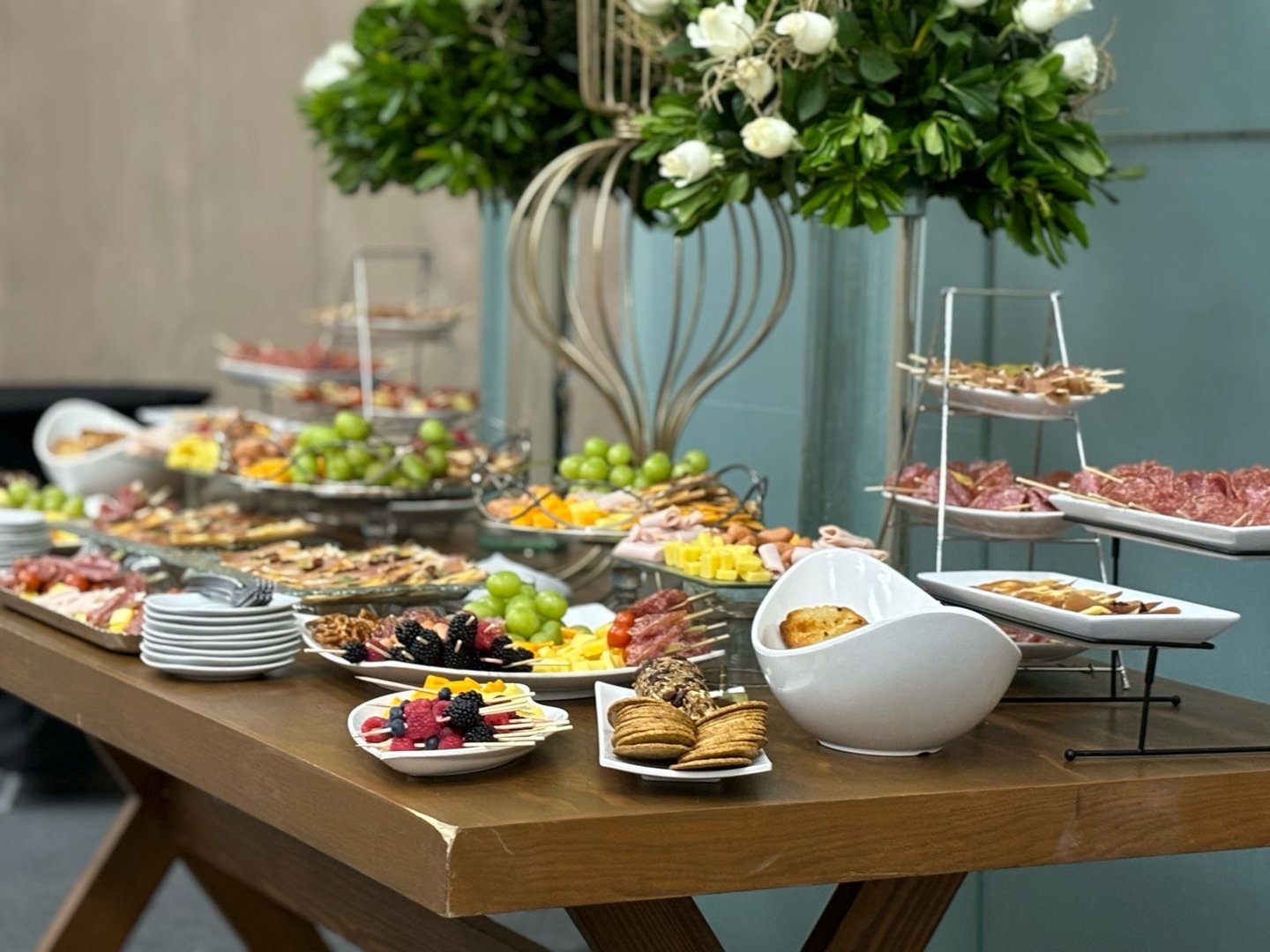 A buffet table with assorted appetizers, fruits, meats, and cheeses, adorned with decorative greenery in the background.