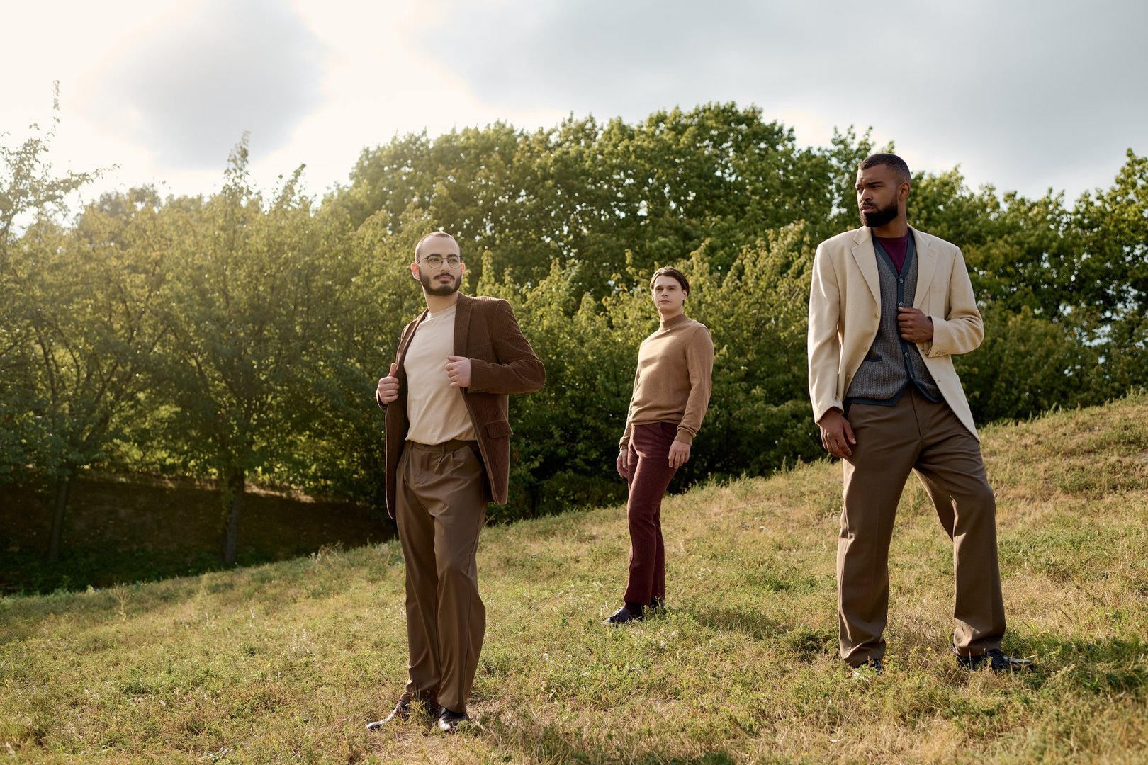 Three fashionable men showcase autumn styles, standing confidently in a beautiful natural landscape.