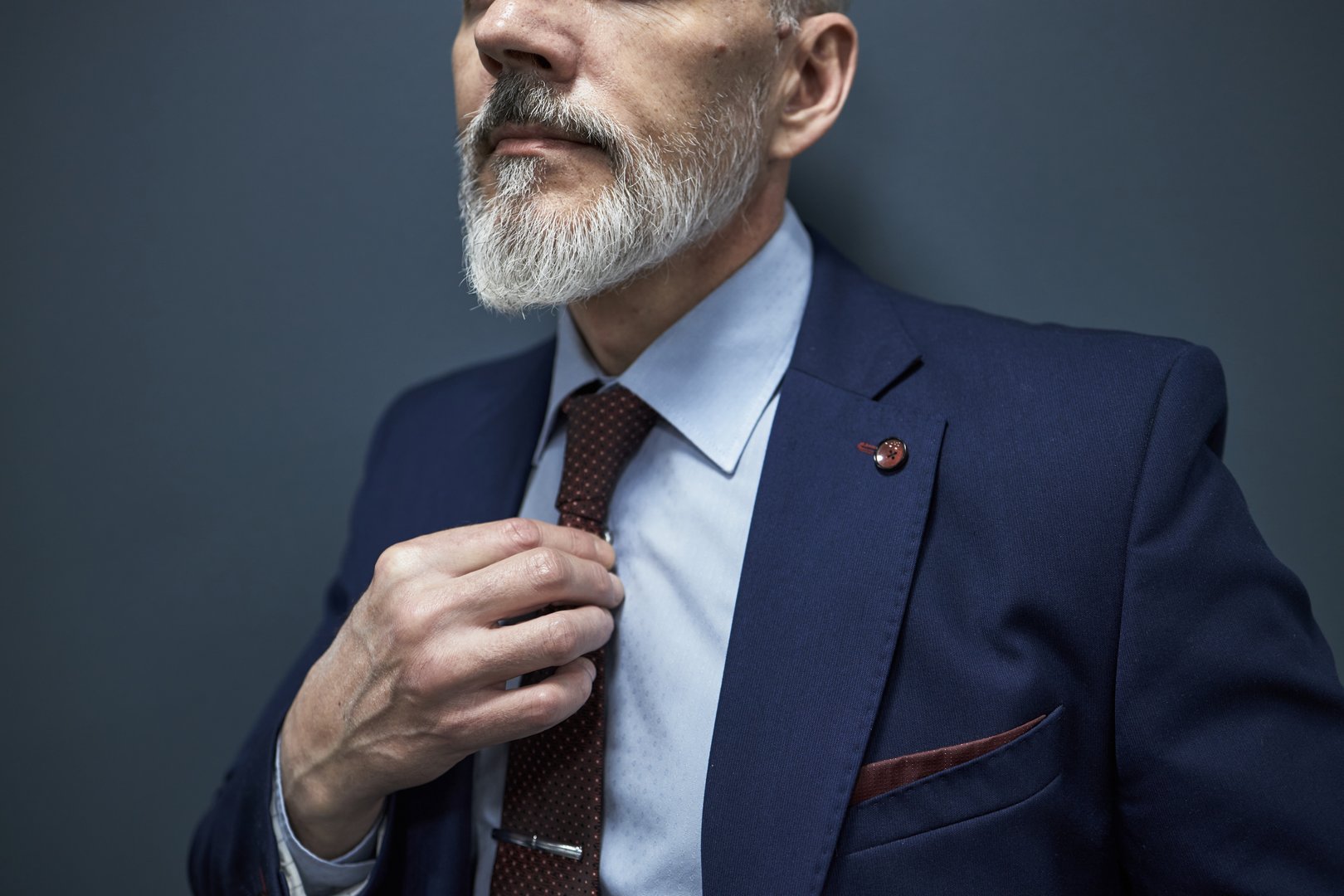 Caucasian senior businessman adjusting tie while wearing formal suit, showing neatly groomed gray beard and mustache, partial face visible, standing against plain blue background