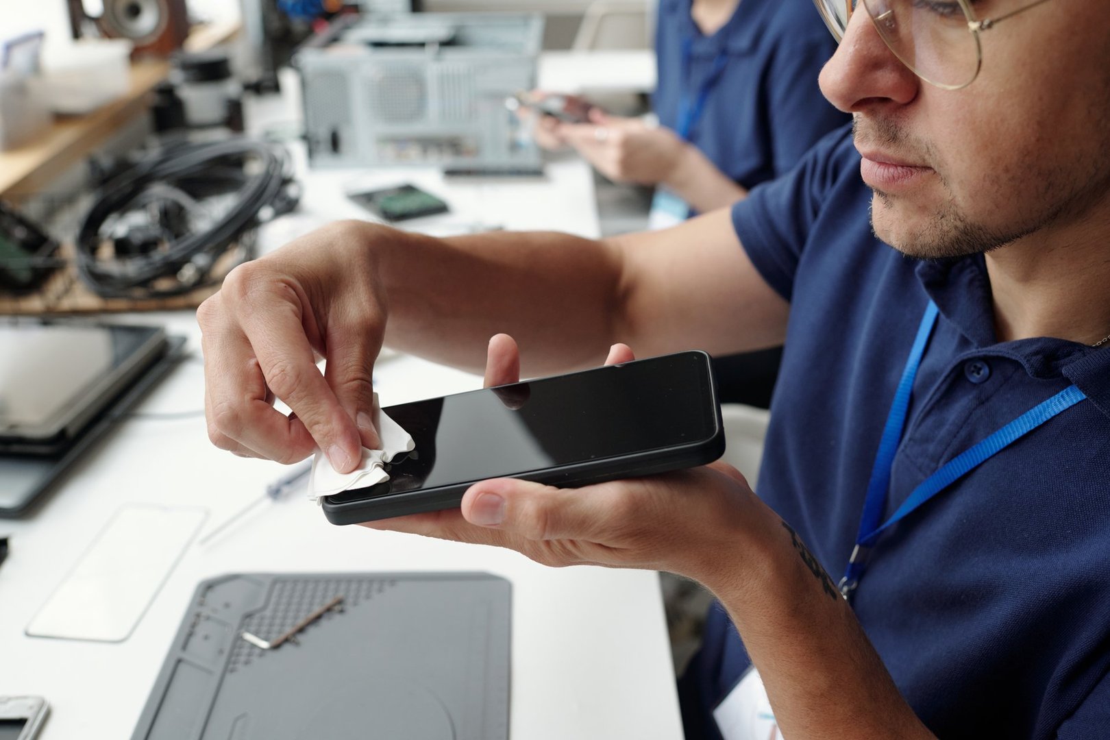 Person carefully cleaning smartphone screen using cloth while sitting at workstation in repair shop surrounded by various tools and devices