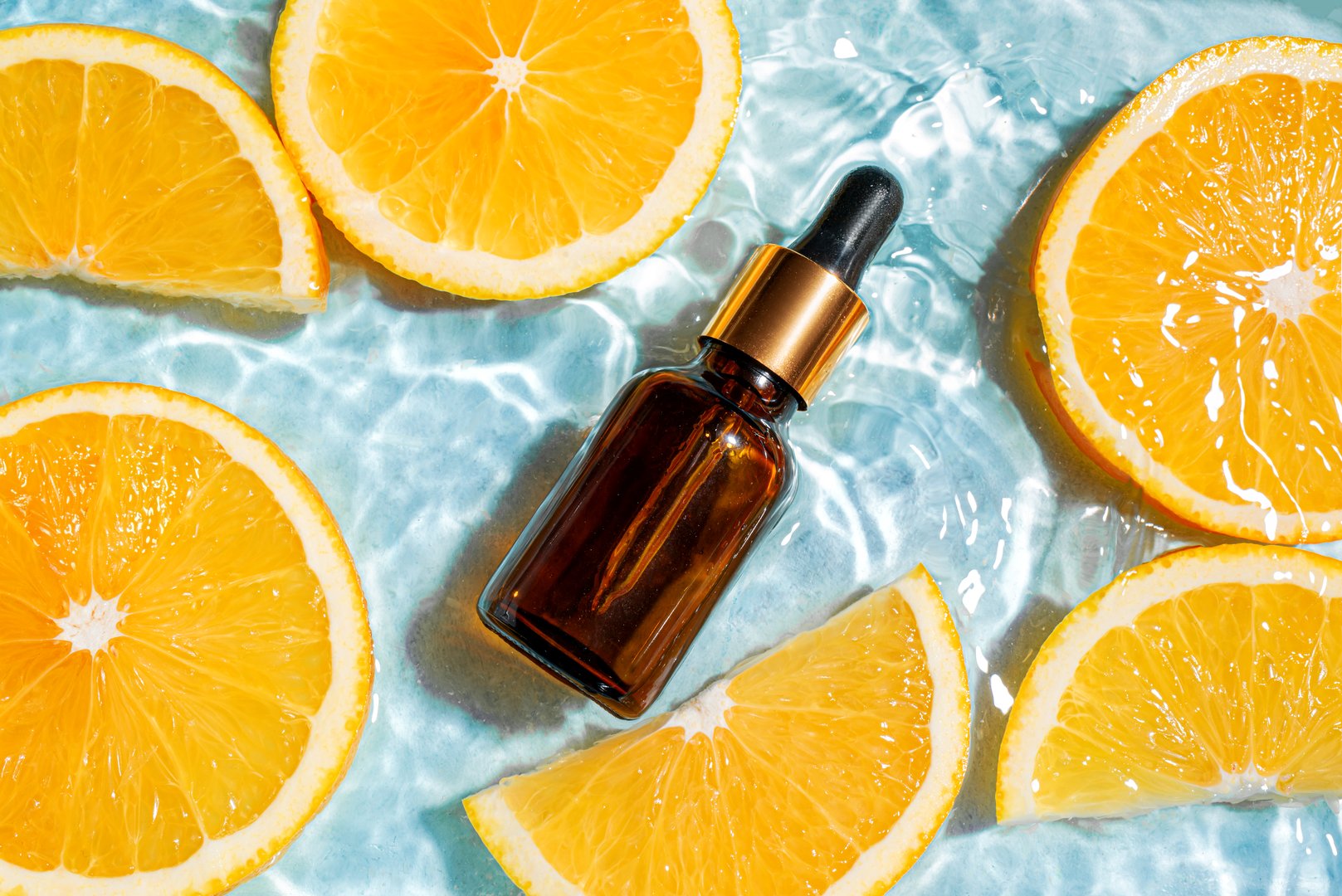 Dropper bottle of cosmetic serum in clear splashing blue water surface with orange slices, top view, copy space. Moisturizing vitamin C or fruit acid beauty product, hydrating face treatment.