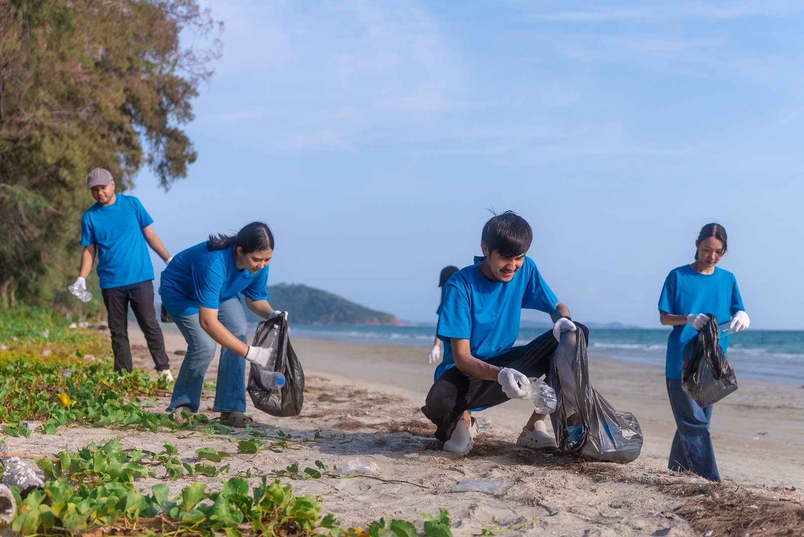 Plastic pollution and environmental problem concept. Happy asian diverse group of volunteers with garbage bags wearing blue t-shirt and cleaning plastic on the beach. Volunteers collecting trash.