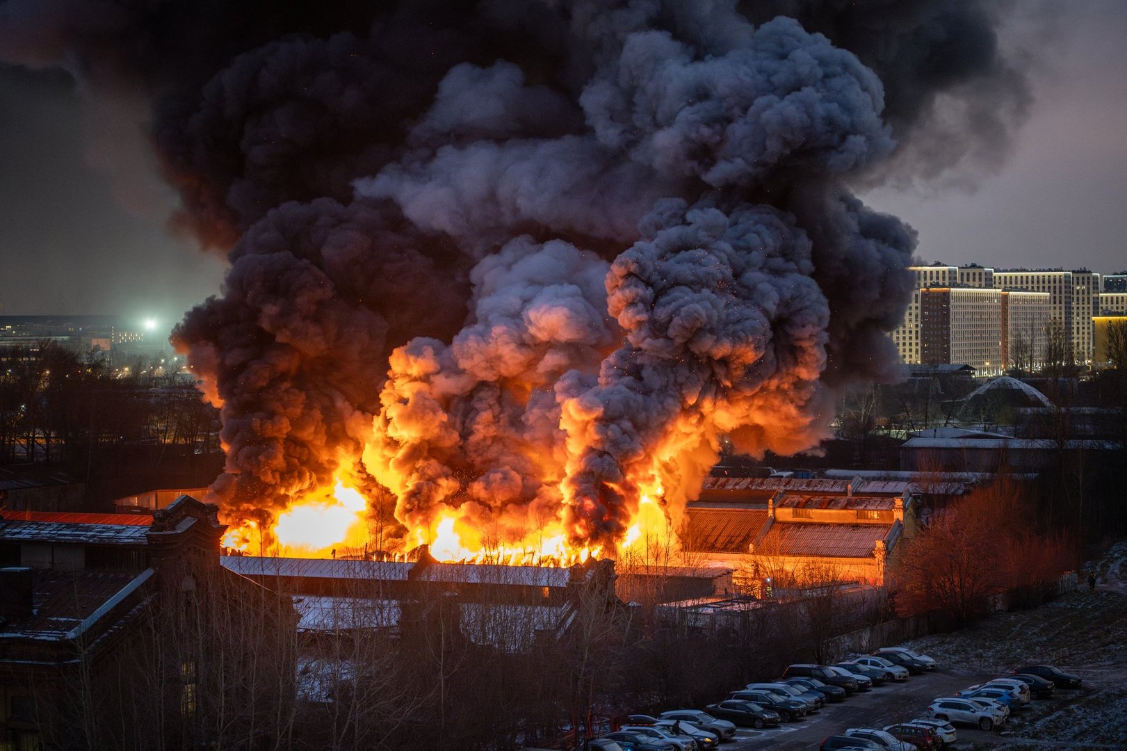 Carelessness with fire hazardous things. Firefighters extinguish house soon collapsing. Nearby burning apartments building with bright orange flames sparks and smoke from strong fire at late night.