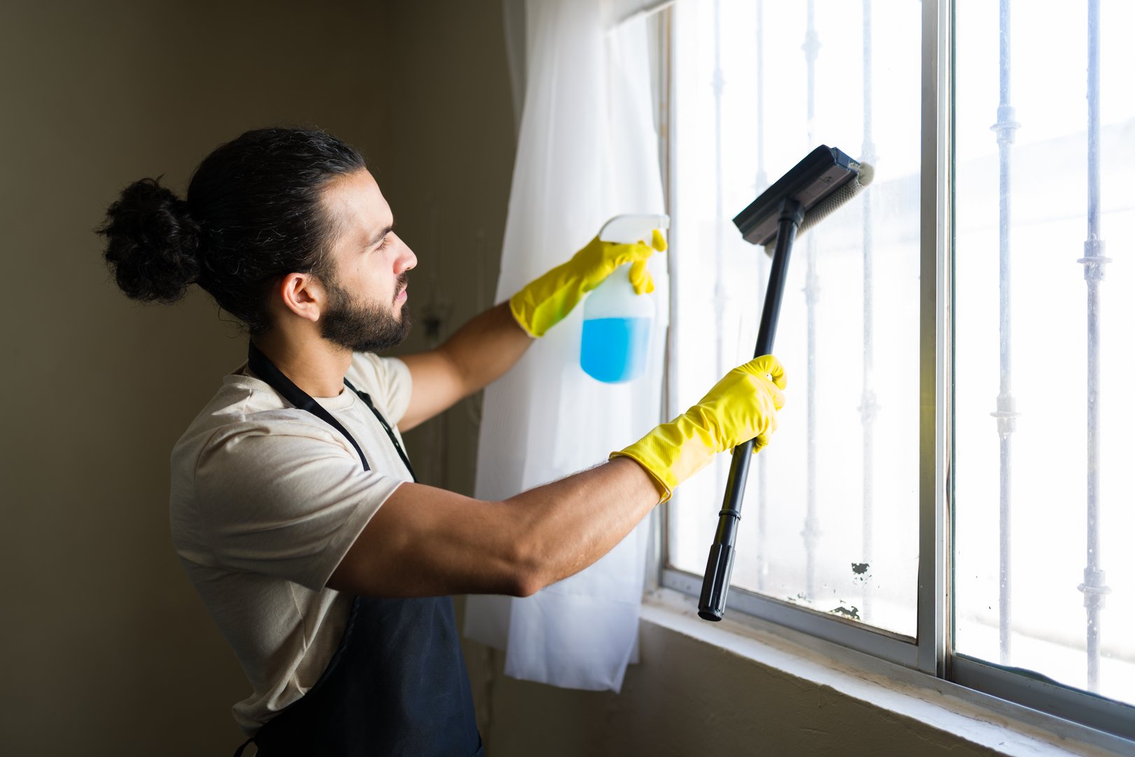 Hispanic cleaner diligently washing windows with cleaning products and gloves, engaging in essential domestic chores for a tidy home