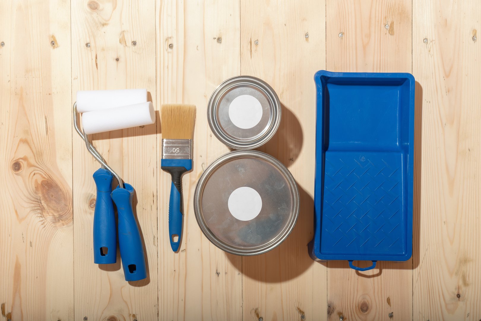 Set of paint rollers, brush, paint cans and blue tray neatly arranged on clean wooden floor, suggesting careful preparation for renovation.