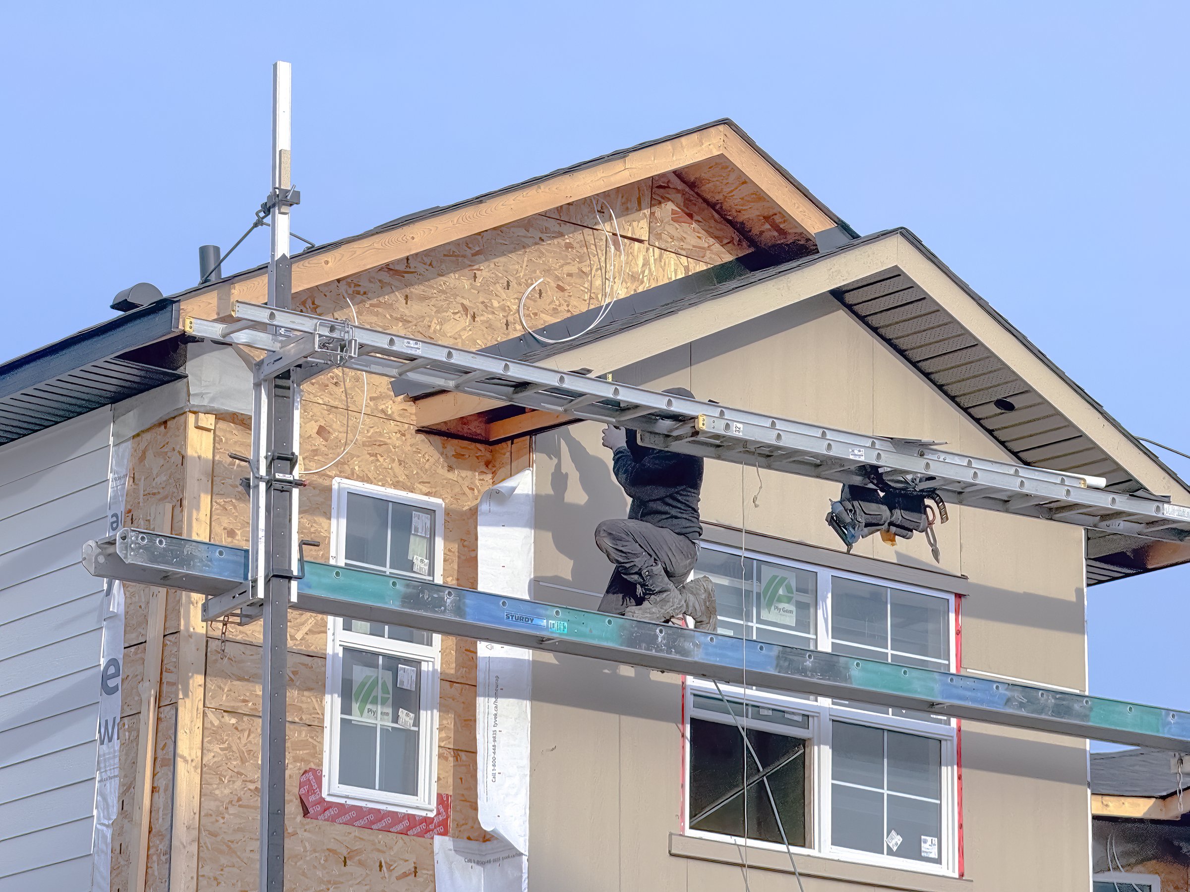 Calgary, Alberta, Canada. Dec 14, 2024. A construction worker carefully maneuvers on scaffolding, installing siding on a new home under construction.