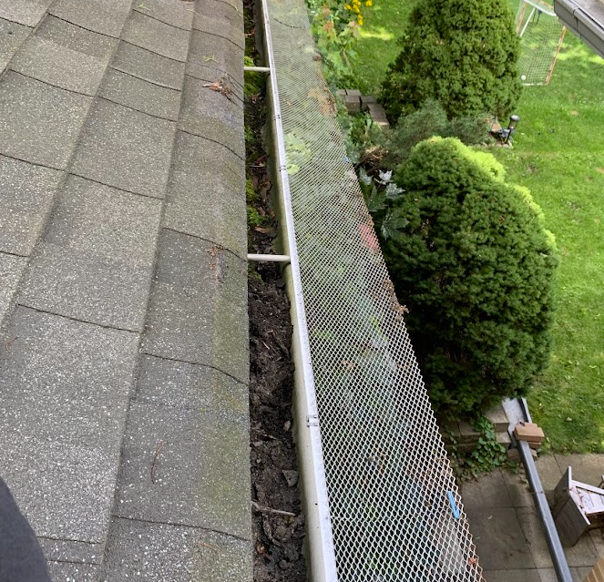A house roof with a gutter guard partially clogged by debris, surrounded by trees and a lawn.