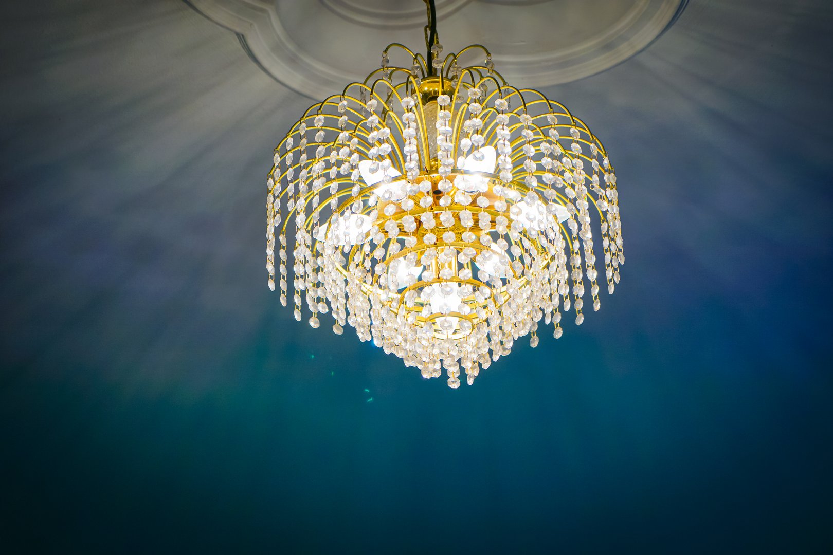 Ornate gold chandelier with crystal prisms hanging from a decorative ceiling, illuminated to cast sparkling light and shadows in a luxurious interior setting.