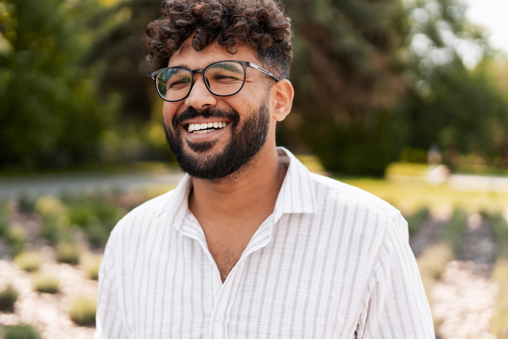 Cheerful young adult with eyeglasses and a beard laughing joyfully in a vibrant park, surrounded by lush greenery and sunlight