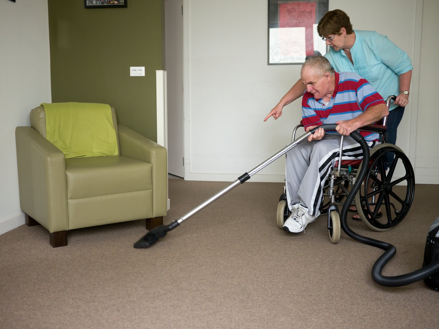 This photo shows a senior man with an intellectual and physical disability vacuuming the floor of his home.  He is being assisted by a home carer.