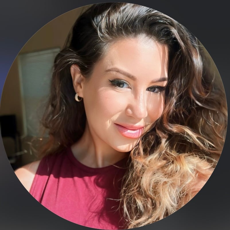 A woman with long wavy hair, wearing hoop earrings and a red top, smiles in natural light.