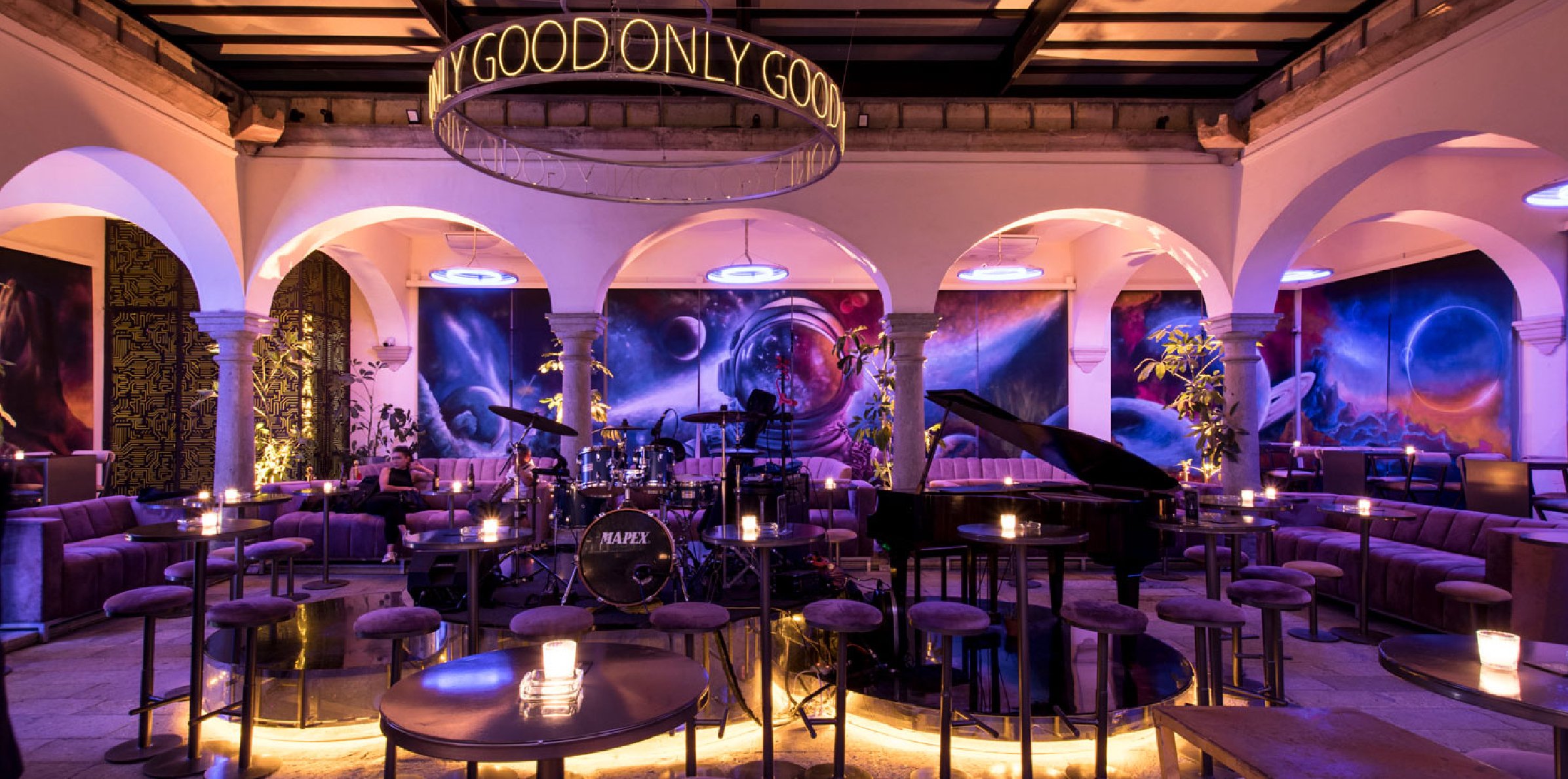 Chic lounge with stage, piano, drums, and ambient lighting. "Only Good" sign above seating area, vibrant art on walls.