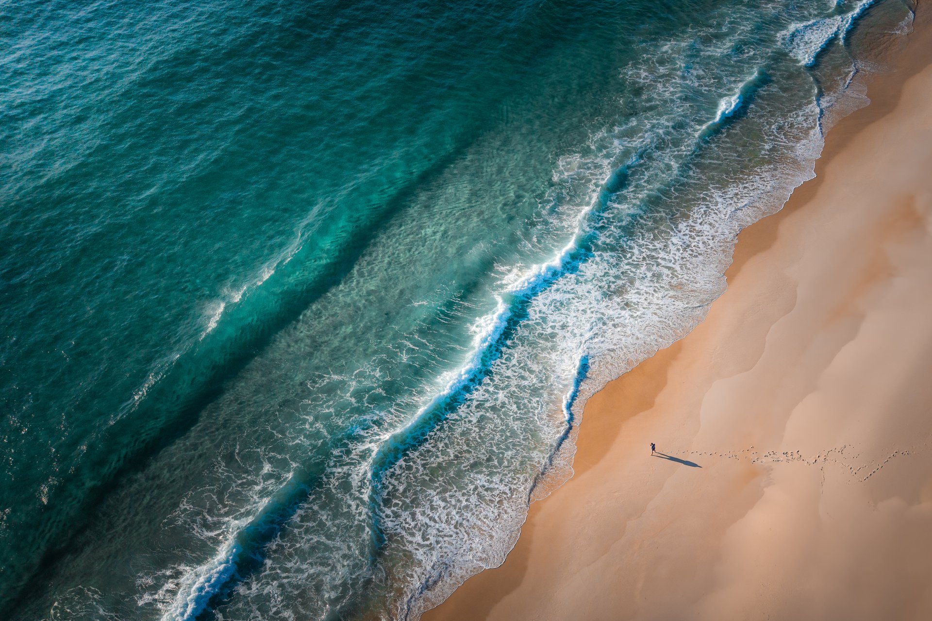 A lone person walks along a remote stretch of golden beach where turquoise waves gently roll onto the shore. Photographed from above, the footprints in the sand and long shadow create a sense of peaceful isolation and connection to nature. Captured on a clear day with vivid coastal colors and texture-rich water.