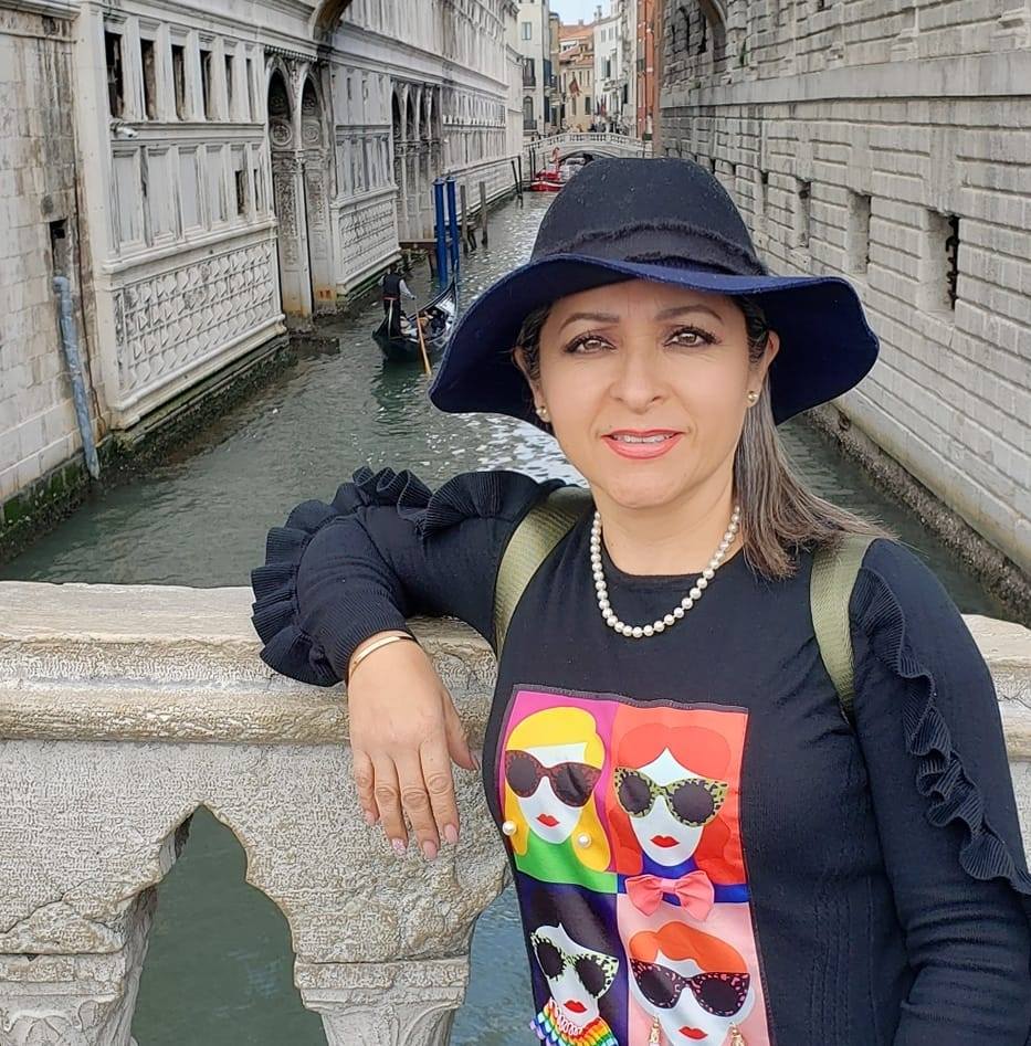 Woman in a black hat posing on a bridge over a canal with gondolas in a historic city.