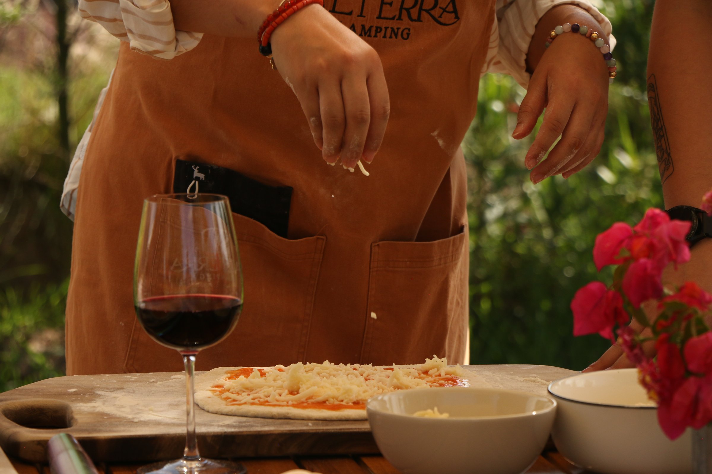 Person in apron preparing pizza with toppings, red wine glass and bowls nearby on a wooden table, flowers in the foreground.