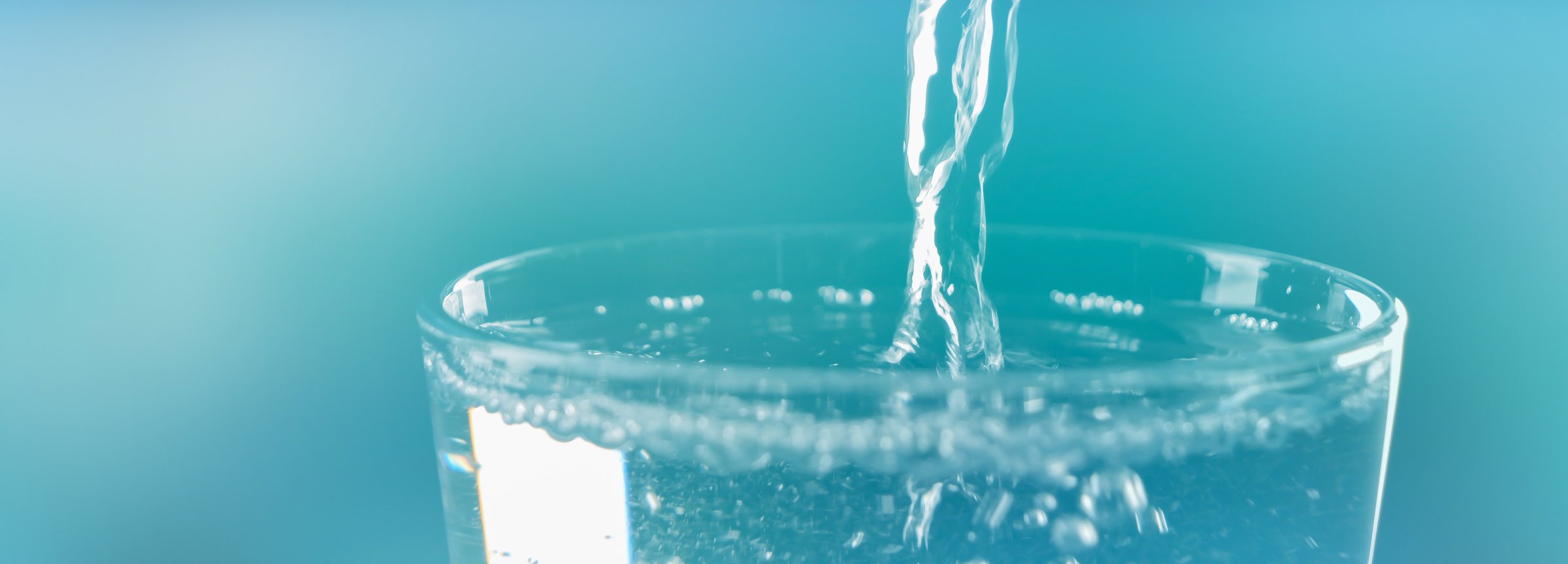 super closeup of pouring a full transparent water glass with water jet isolated on blue background banner with sunlight