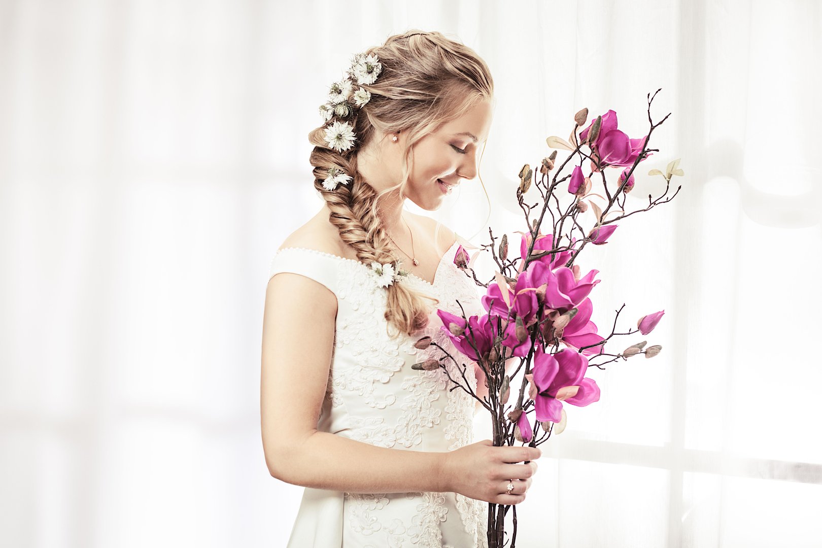 A portrait of a blond smiling bride with a bouquet of magnolias by curtains