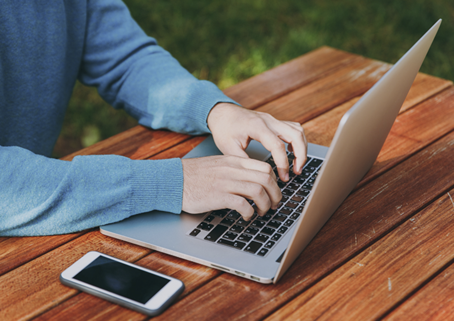 Person typing on a laptop at a wooden table outdoors, with a smartphone placed beside the laptop.