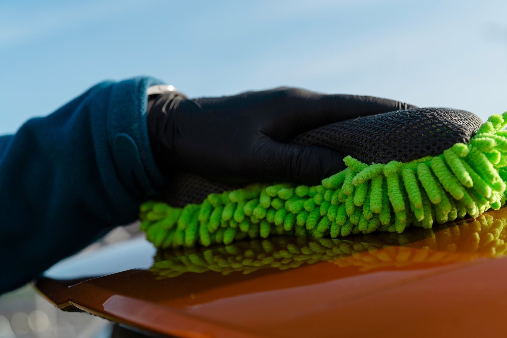 A man wearing a blue jacket and black gloves is wiping and polishing surface of orange car with yellow microfiber