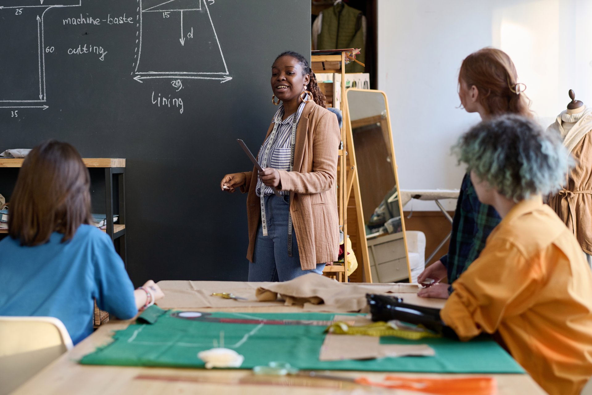 Portrait of African American female teacher standing by board explaining clothing making to group of girls in atelier workshop copy space