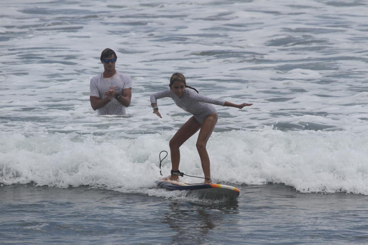 A young person surfing a small wave on a surfboard while an instructor watches from the water.