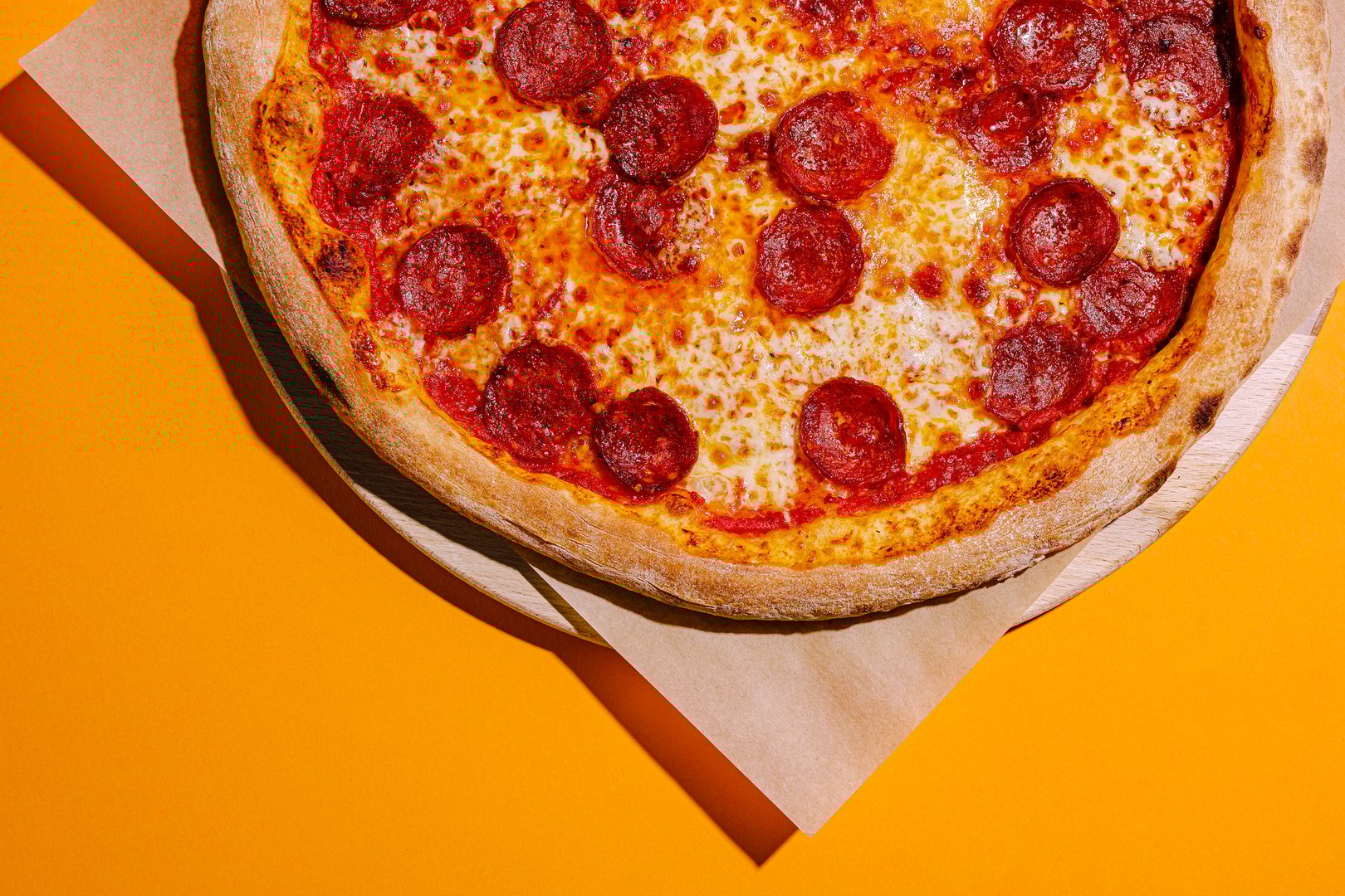 A freshly baked pepperoni pizza rests on a wooden board against a bright orange backdrop, showcasing its cheesy topping and crispy crust, ready to be enjoyed.