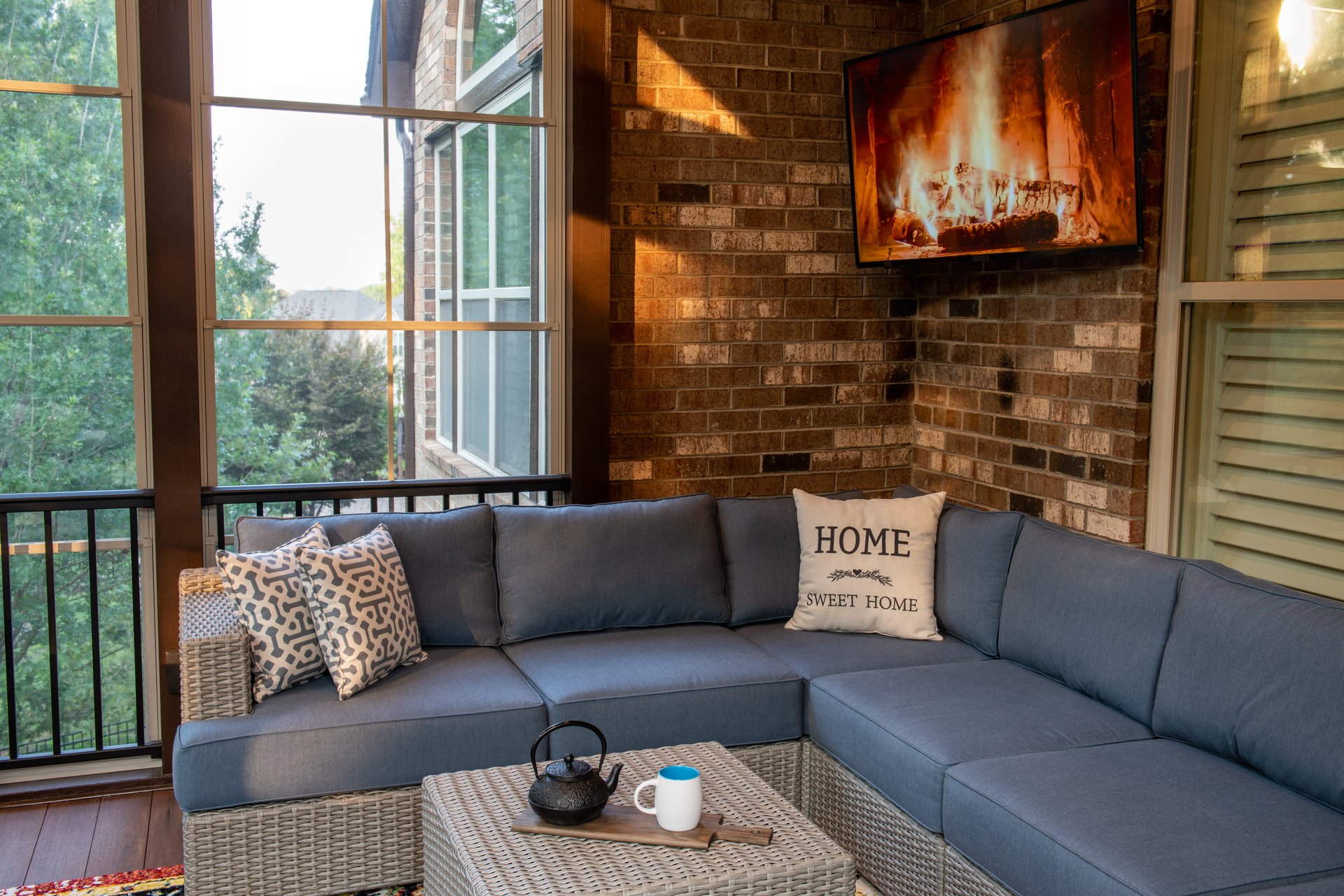 A cozy corner of an outdoor living space with modern porch windows, patio furniture, cushions and pillows. Coffee or tea pot and cups on coffee table and TV mounted on brickwall.