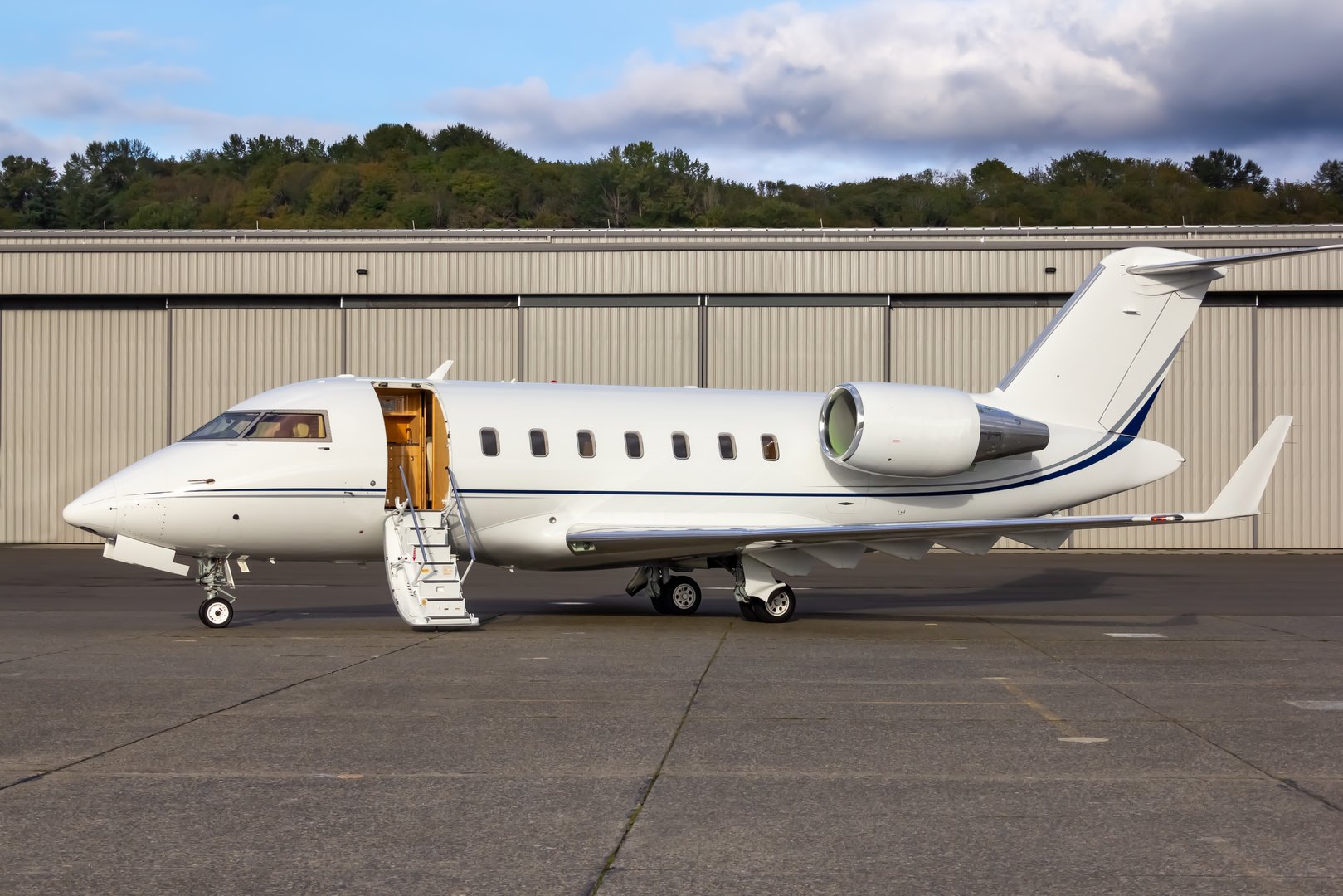 The image shows a Bombardier Challenger 650 corporate business jet aircraft originally developed by Canadair.  The aircraft are now being produced by Bombardier and is still in production.  The jet is parked on the ramp in front of a hangar with the entry door open and stairway down.  The photo evokes a feeling of  importance and wealth.  The photo has blue sky with a few clouds.  It is late in the day adding a warm tone to the image.