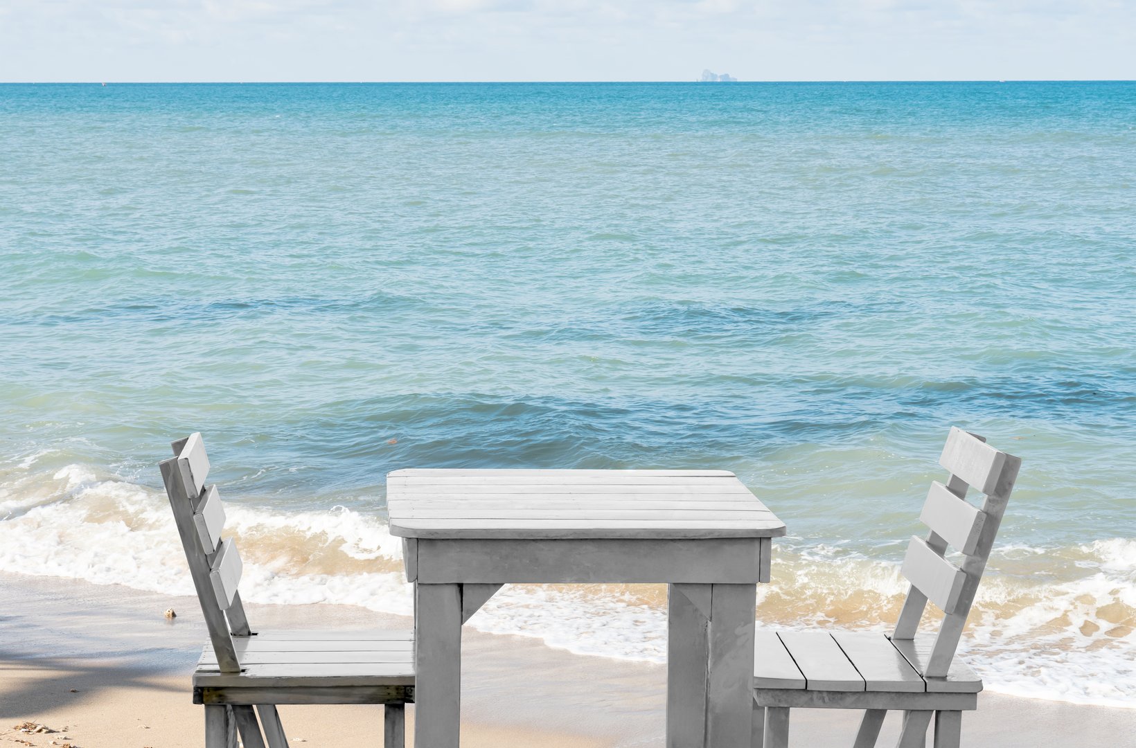 Empty White wooden table and chairs on tropical sand beach with sea wave,blurry sky background. Image summer vacation concept for travel, relaxation, romantic dining, or coastal lifestyle design.