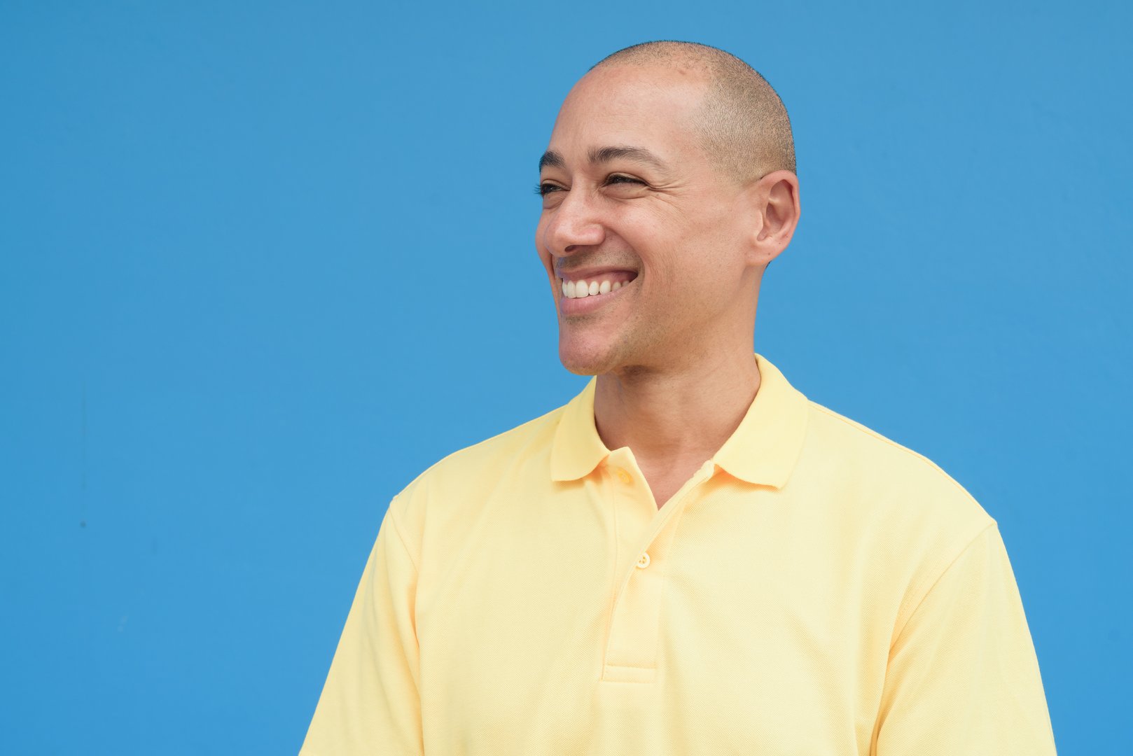 Studio portrait of a handsome mature Italian man with shaved head wearing yellow polo shirt, standing in front of a neutral plain blue wall with copy space. Clean isolated cut out look.