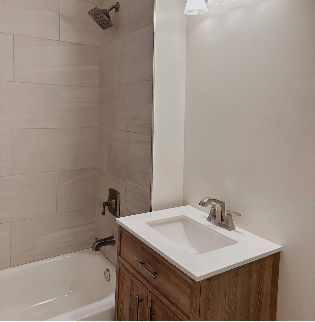 Modern bathroom with wood vanity, white sink, and tiled bathtub shower combination with metal fixtures.