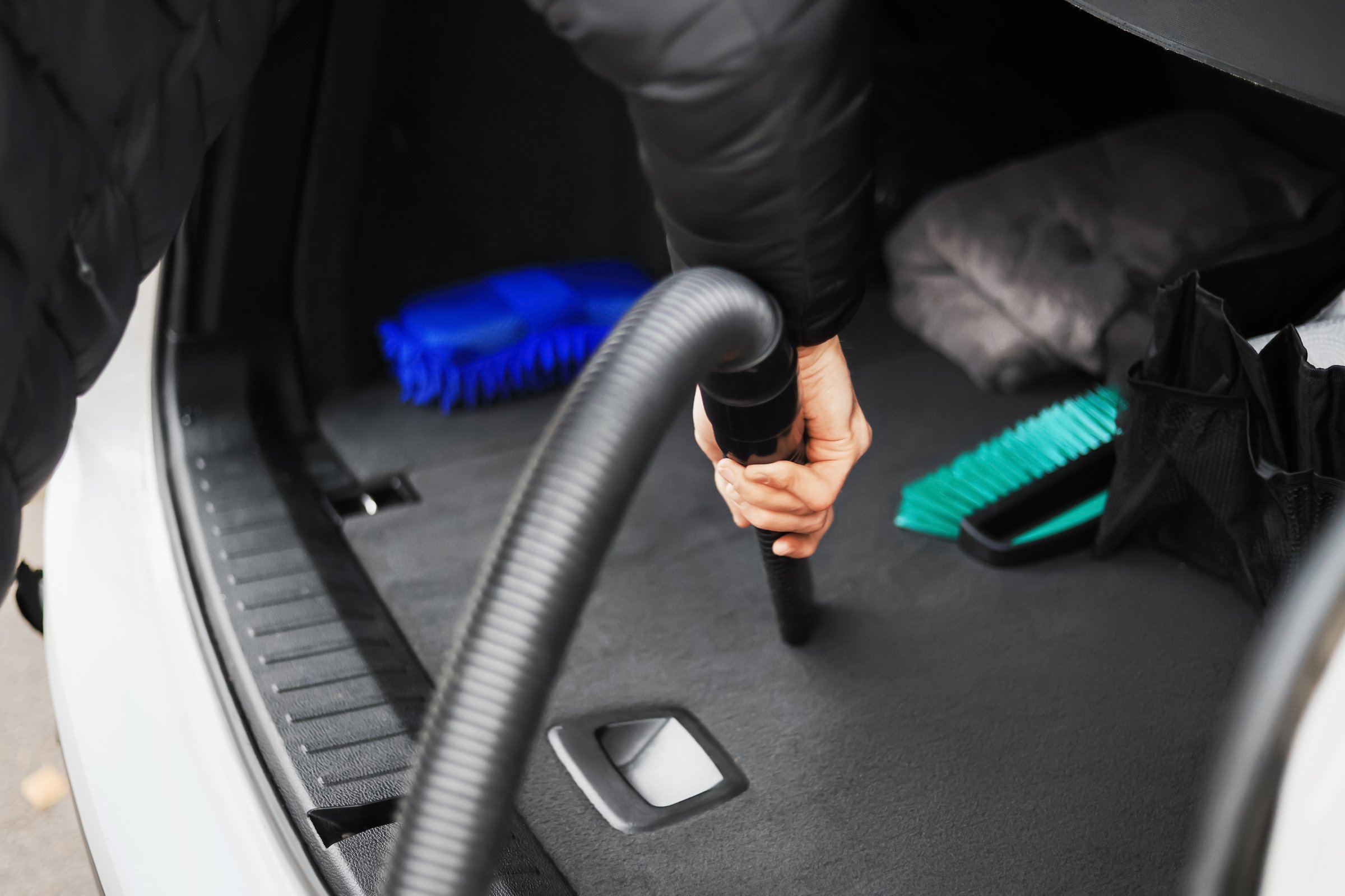 The man vacuums the interior of a car at a self-service car wash. The scene includes cleaning tools and a vacuum hose.