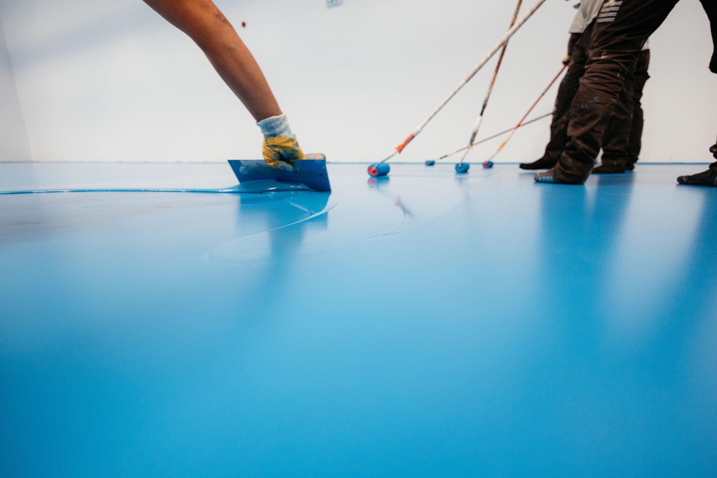 Workers applying a blue epoxy resin on a floor with rollers and spatulas in an industrial setting.