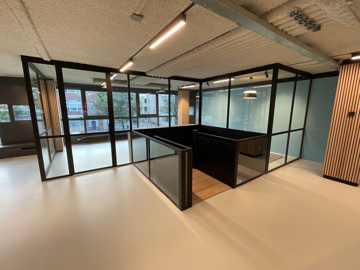 Modern office space with glass walls, wooden accents, and overhead lighting, featuring large windows and clean design.