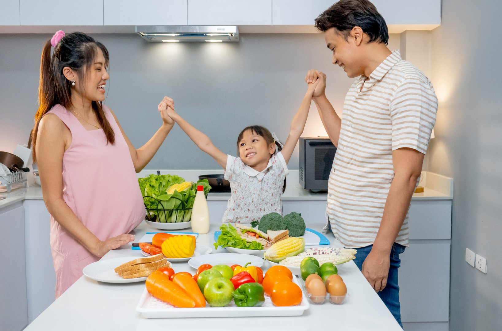 Little Asian girl hold hands together with father and pregnant mother and stay in kitchen to prepare for cooking with happiness.