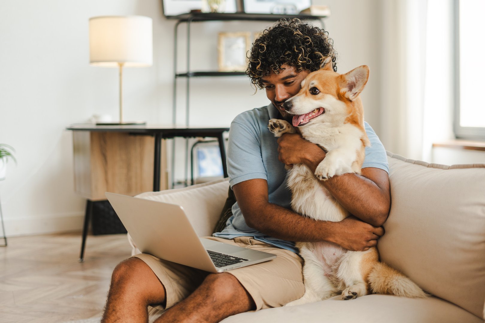 Attractive Indian man, freelancer working remotely from home and hugging his corgi dog using laptop, watching video. Online technology concept