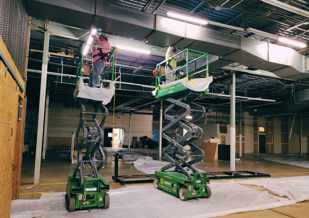 Workers on scissor lifts performing maintenance in a large industrial facility with plastic sheets covering the floor.