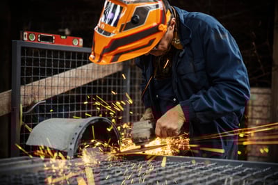 Male industrial worker wearing protective mask and uniform using an angle grinder to cut metal, creating many sparks