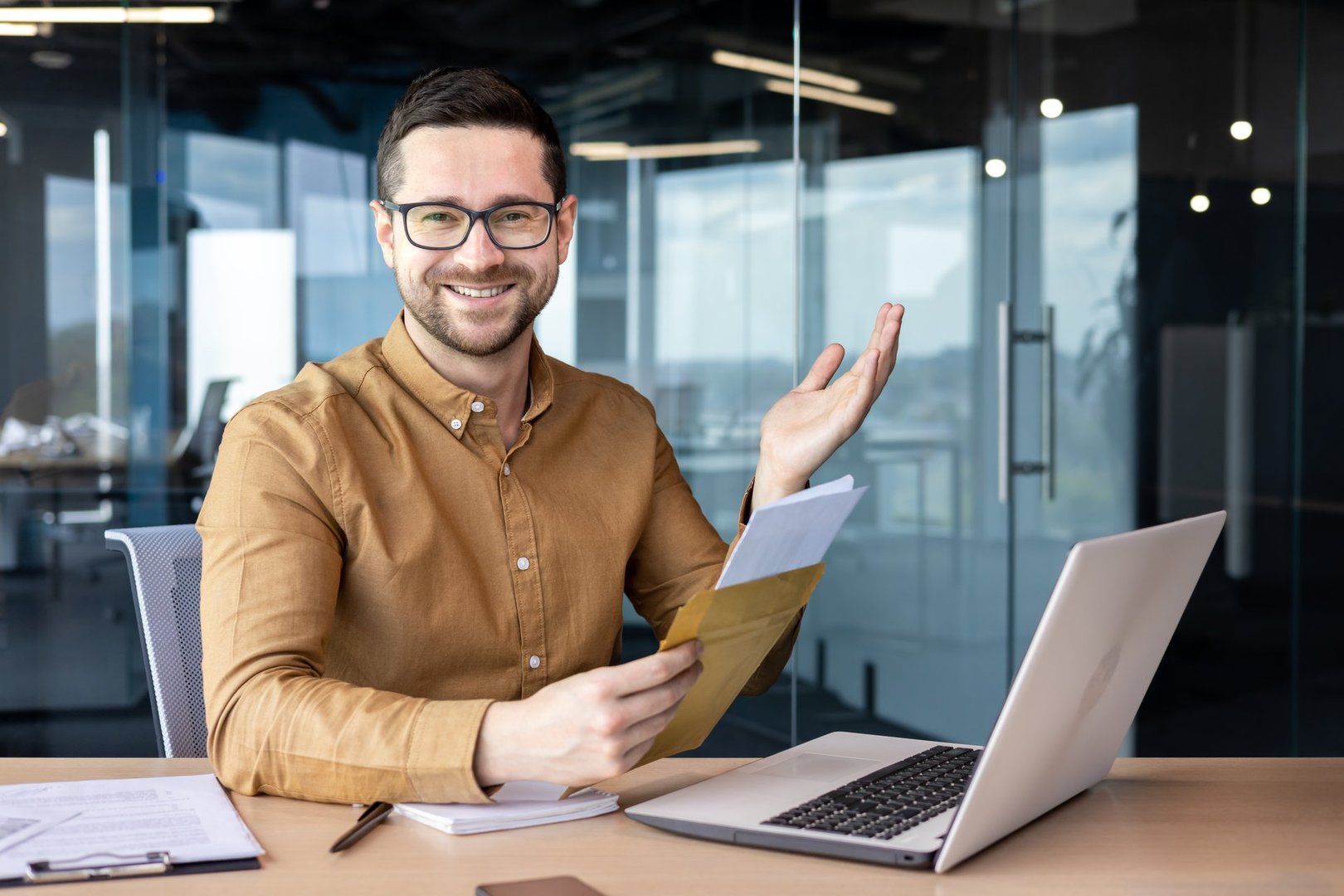 Portrait of a man sitting in the office and working on a laptop. Received and holds in his hands an envelope with a letter and documents. Smiling looking at the camera.