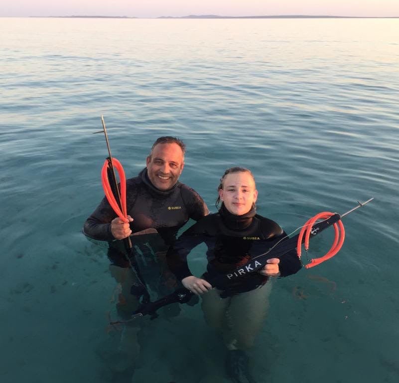 Two people in wetsuits standing in calm water, holding spearguns, with a clear sky in the background.