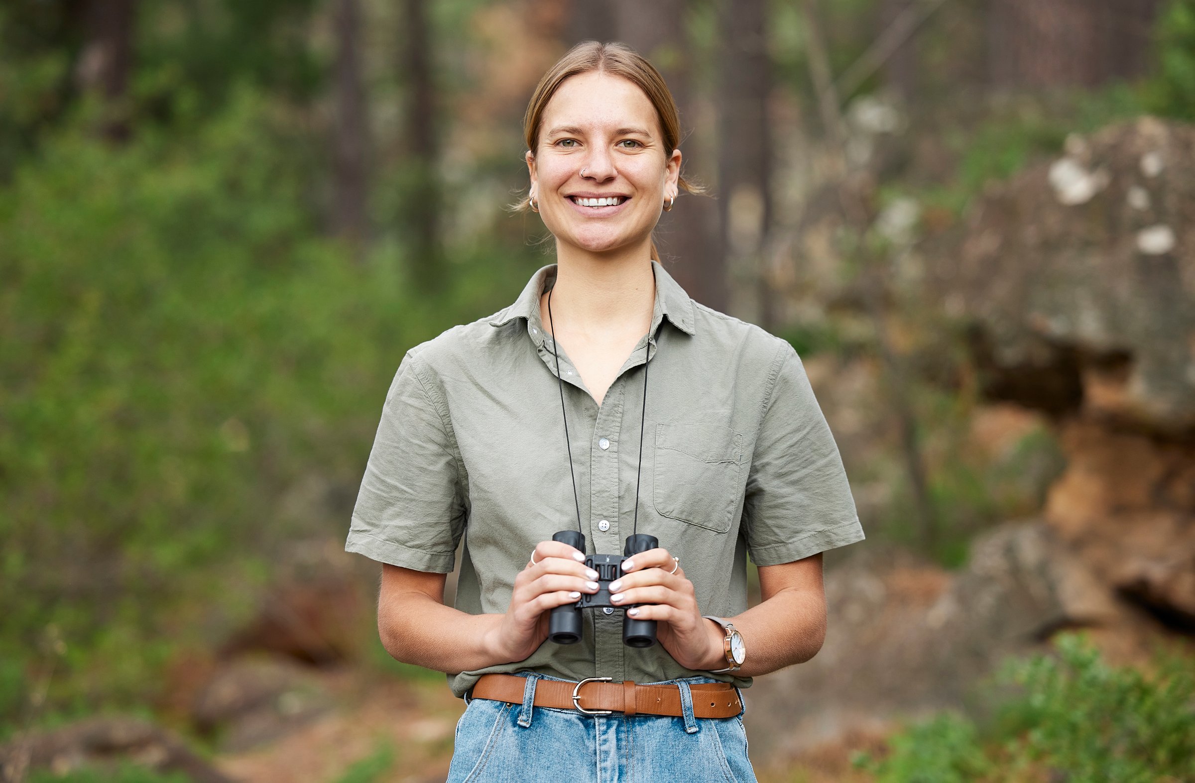 Binocular, forest and portrait of happy woman hiking for nature journey, jungle adventure and travel or outdoor explore. Face of a young person birdwatching and trekking in eco friendly, green woods