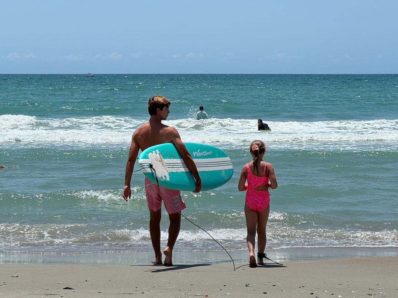 Man with a surfboard and a girl walk towards the ocean on a sandy beach under a clear blue sky.