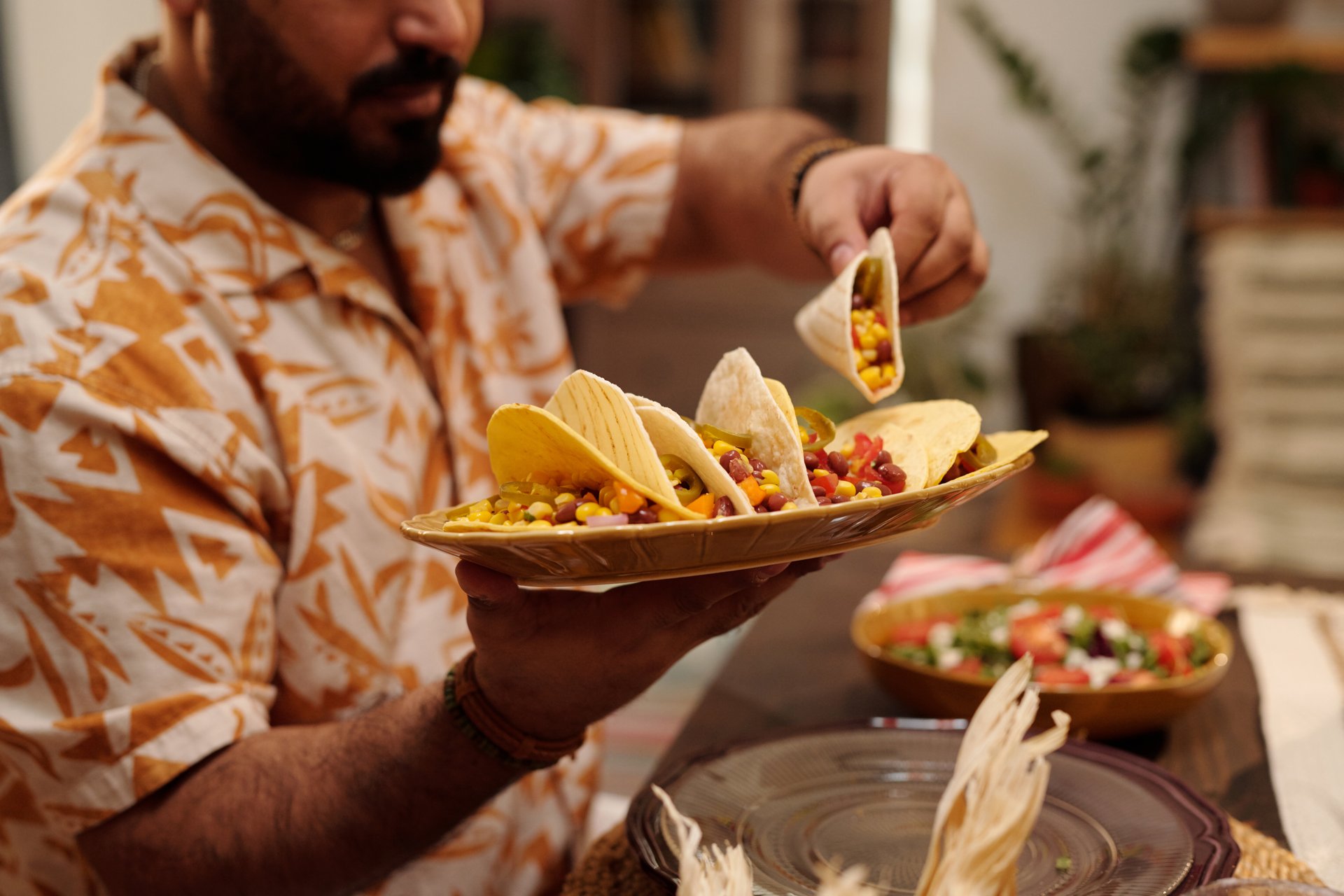 Hands of young Hispanic man holding plate with tasty Mexican tacos and taking one of them while sitting by served festive table at dinner