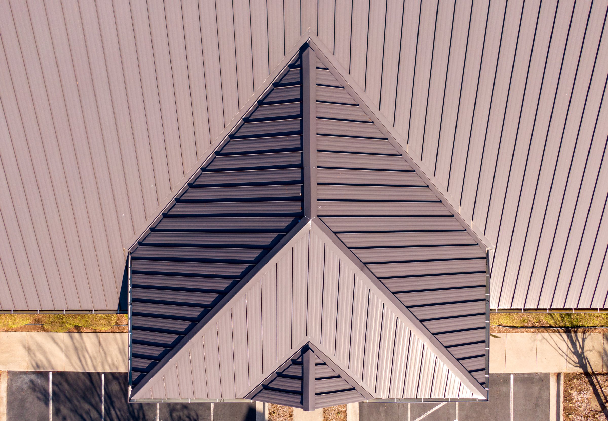 An elevated, high-resolution drone shot showcasing the sleek, standing seam metal roof of a modern commercial building. The image highlights the clean lines, sharp angles, and durable, dark gray finish of the metal roofing system, which provides superior weather resistance and a contemporary architectural look.