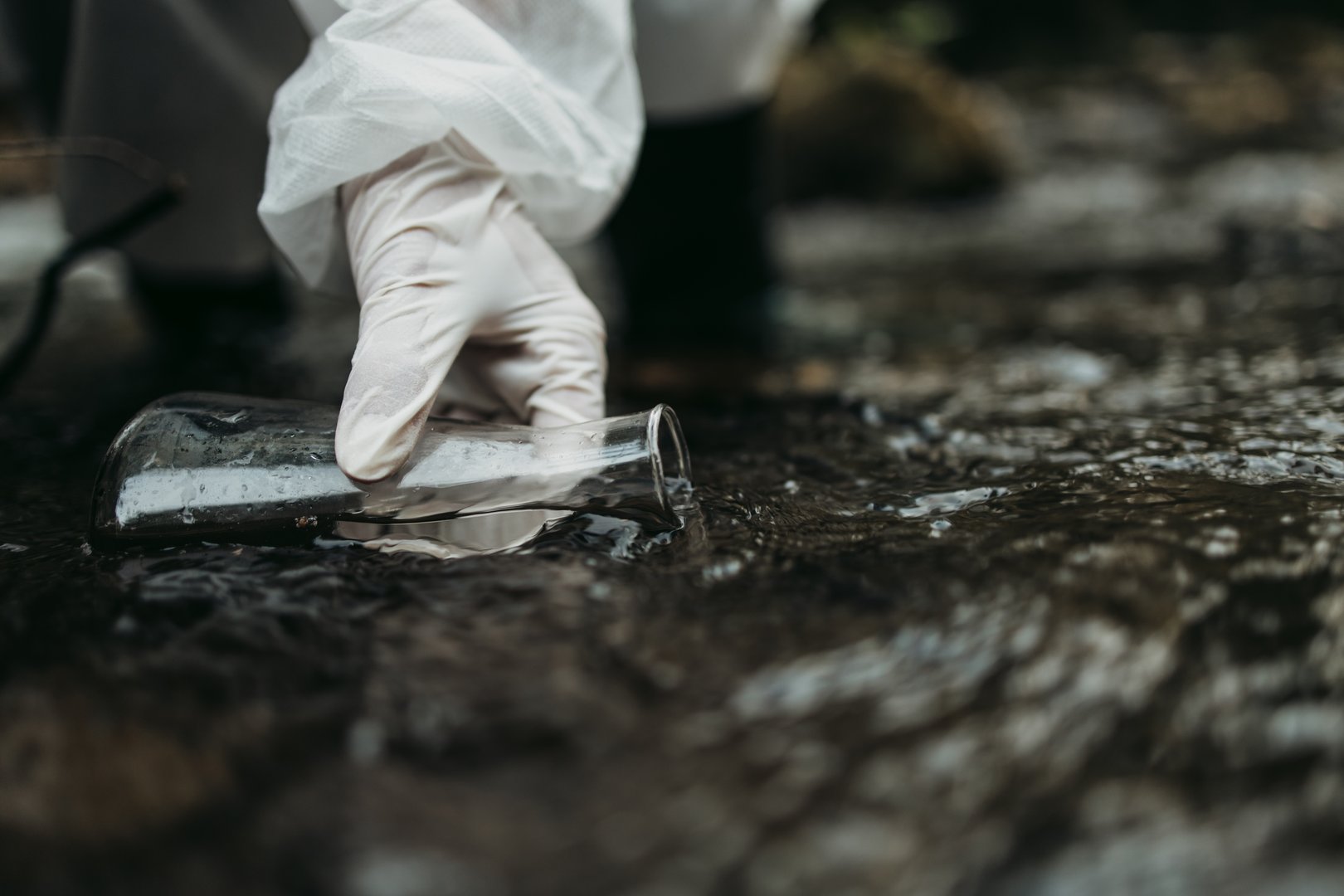 Close up shot of scientist biologist and researcher in protective suit taking water samples from polluted river