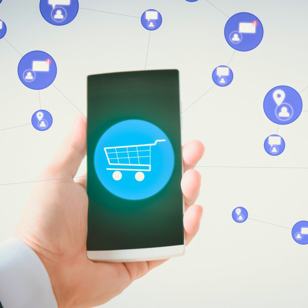A hand holds a smartphone displaying a glowing shopping cart icon. Social media notification bubbles float around the device.